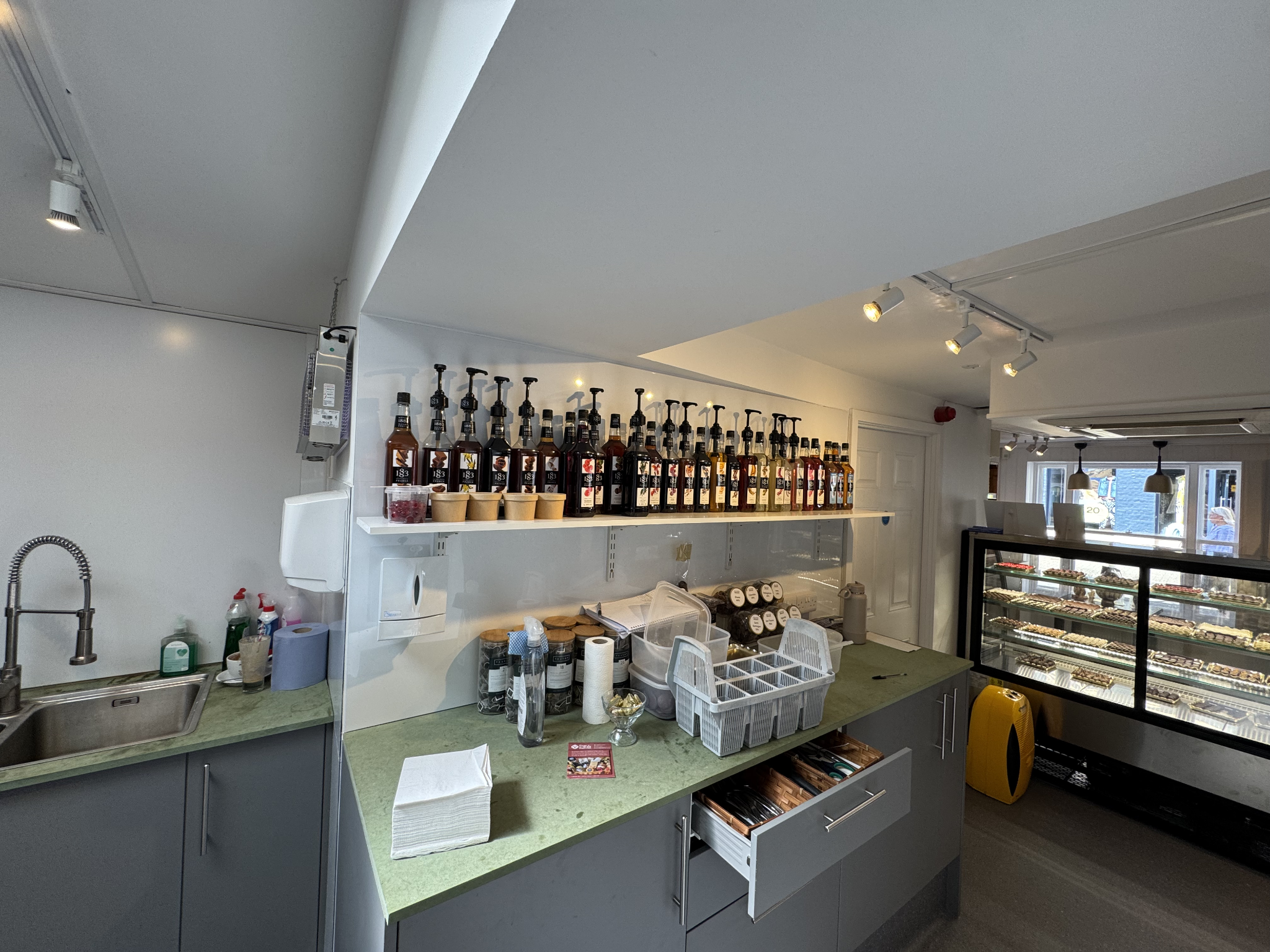 Kitchen prep area with stainless sink, green countertop and shelf of flavoured syrups